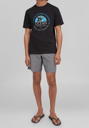 T-shirt en coton noir avec un graphisme circulaire "Our Ocean Mission" en bleu et blanc. Associé à un short gris et des tongs noires.