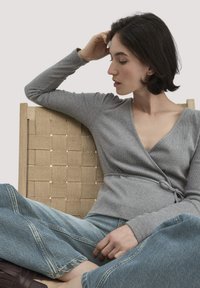 Grau umwickelter Pullover aus gerippter Textur mit V-Ausschnitt und tailliertem Bund. Kombiniert mit hellblauen, weit geschnittenen Jeans, sitzend auf einem geflochtenen Stuhl.