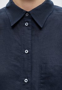 Κοντινό πλάνο ενός ατόμου που φοράει σκούρο μπλε πουκάμισο με γιακά και τσέπη στο στήθος, με τέσσερα εμφανή κουμπιά κουμπωμένα.