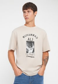 Bežs kokvilnas T-krekls ar melnu grafiku, kurā attēlots Muhameda Ali, ar tekstu "Muhameds Ali – tautas čempions" kontrastējošās krāsās.
