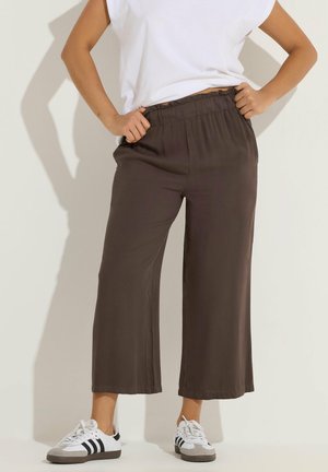 Persona che indossa pantaloni marroni a gamba larga con vita elastica e sneakers bianche con strisce nere, mani che tengono la vita.