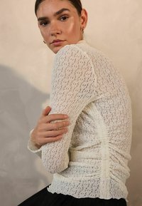 Light cream, textured top with a fitted design, featuring gathered details along the sides and delicate lace-like patterns. Long sleeves.