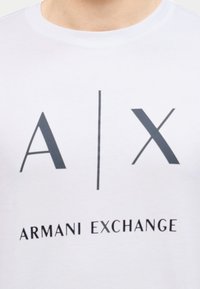 Κοντινό πλάνο ενός λευκού πουκάμισου με μαύρα γράμματα "A | X" και το κείμενο "ARMANI EXCHANGE" κάτω από το λογότυπο στο στήθος.