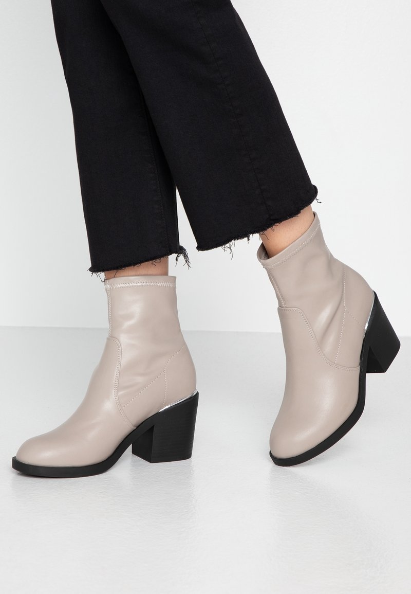 Bottines beige à talons blocs portées avec un pantalon court noir sur un sol et un fond blancs.