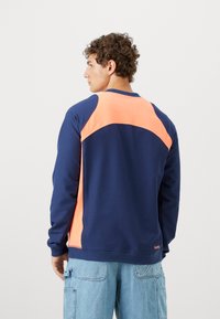 Mørkeblå sweatshirt med et klart orange panel hen over ryggen og skuldrene. Har lange ærmer og rund halsudskæring.