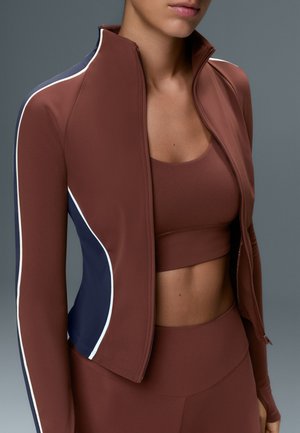 Femme portant une veste marron zippée avec des bandes latérales bleu marine et blanches sur un brassière de sport marron et un legging taille haute assorti.