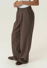 Pantaloni marroni a vita alta e gamba larga realizzati in tessuto liscio con pieghe nette e tasche laterali. Combinati con scarpe scure.