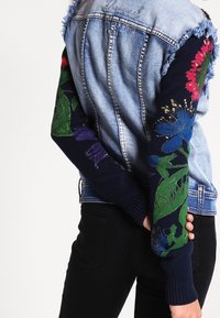 Jeansjacka med fransade kanter och broderade blommormönster på ärmarna i marinblått och olika klara färger, med en figursydd stil och subtil logotypdetalj.