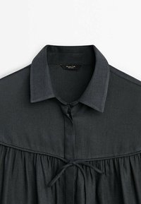 Zwarte button-up blouse met een wijdvallende kraag, textuurstof en geplooide bovendel. Heeft een strik in de taille en een label op de nek.
