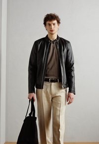 Chaqueta de cuero negra con un cuello alto, camiseta marrón debajo, pantalones beige claros, sosteniendo un bolso negro de tela. Textura suave.