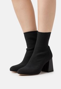 Botas negras hasta el tobillo hechas de una tela texturizada, con un tacón cuadrado y un diseño ajustado que se adapta suavemente al tobillo. Punta redondeada.