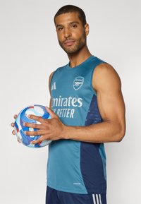 T-shirt de sport sans manches teal avec des accents bleu foncé et le texte "Emirates Fly Better" ; joueur tenant un ballon de football multicolore.