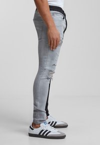 Jeans ajustados gris claro con roturas desgastadas, que presentan un panel lateral oscuro y una cinturilla con cordón, combinados con zapatillas blancas y negras.