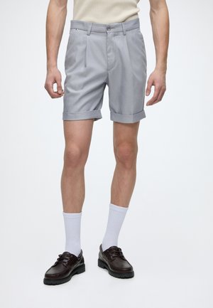 Man wearing light gray cuffed shorts, white crew socks, and dark brown leather lace-up shoes, standing against a plain background.