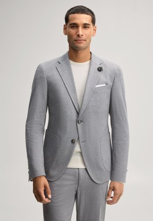 Junger Mann trägt einen hellgrauen Anzug mit weißem Hemd und einer kleinen Blumen-Anstecknadel am Blazer, stehend vor einem einfarbigen Hintergrund.