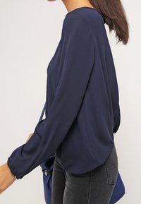 Námořnická modrá halenka s dlouhým rukávem vyrobená z lehkého materiálu, s kulatým výstřihem a volným střihem, nošena s tmavými džínami a křížovou taškou.