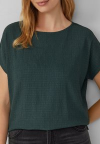 Tegengroen T-shirt met korte mouwen en een gestructureerd, geribbeld oppervlak; ronde halslijn; relaxte pasvorm. Gecombineerd met donkere jeans voor contrast.