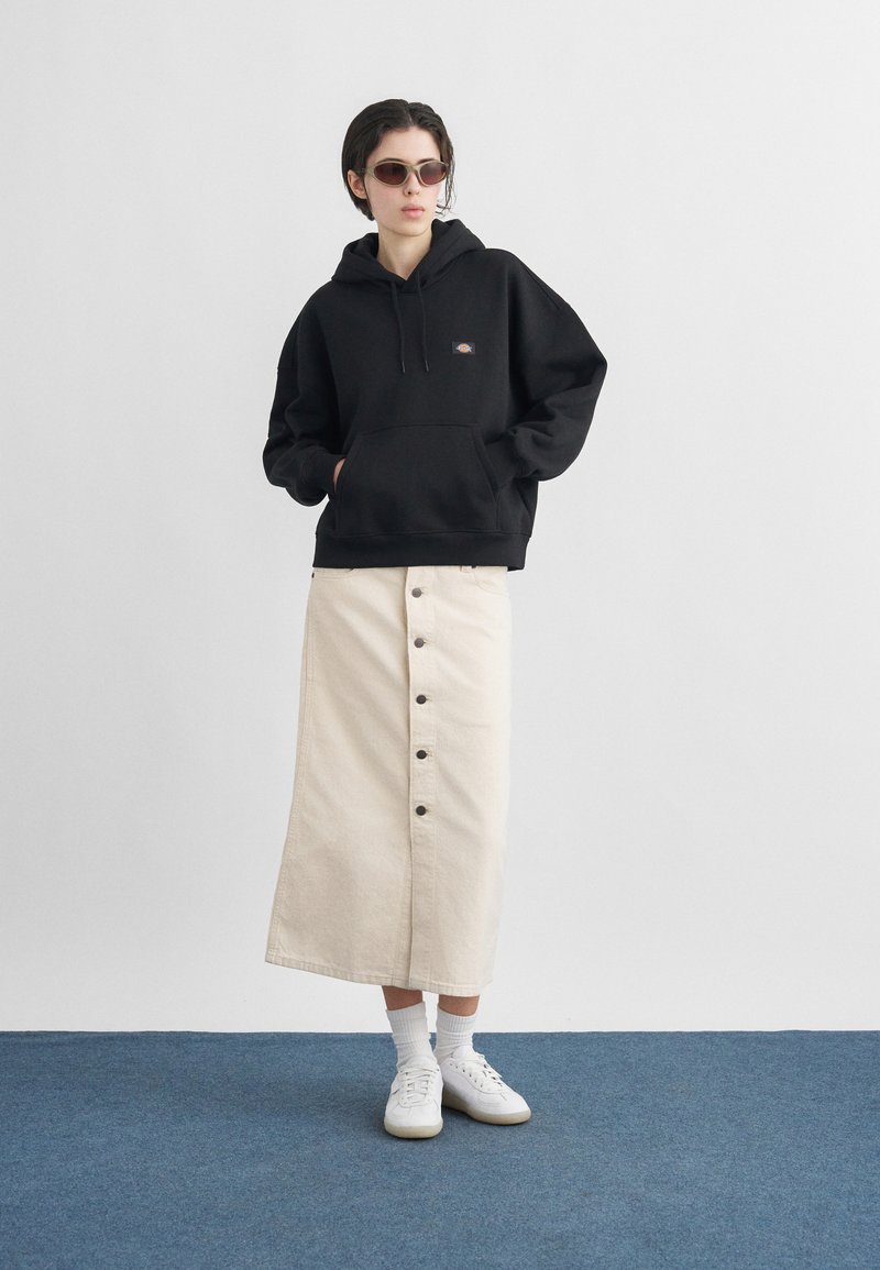 Person wearing black hoodie, beige buttoned midi skirt, white socks, white sneakers, and sunglasses standing on blue floor against white wall.