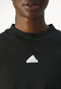 Maglietta nera in cotone con colletto a costine e una toppa con il logo bianco di Adidas sul petto sinistro. Il tessuto appare liscio e leggero.