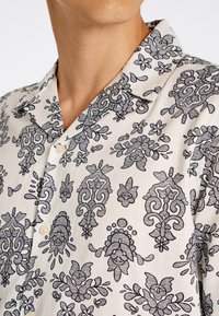 Camisa de manga corta en tela crema con un detallado patrón floral y ornamental en negro, con cuello y cierre de botones.