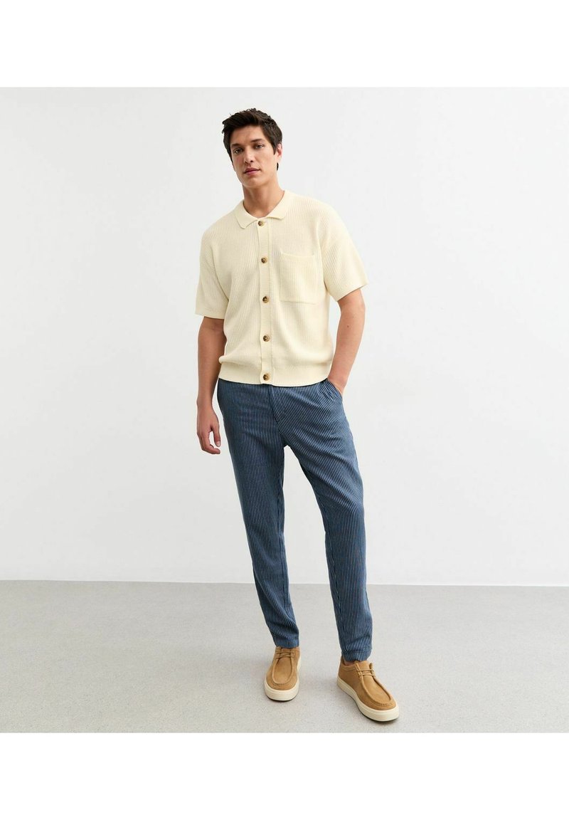 Korte mouwen ribbed shirt in crème met houten knopen, gecombineerd met blauw gestreepte broeken en lichtbruine instapchoenen.