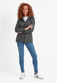 Woman with curly red hair standing with arms crossed, wearing a zip-up star-patterned jacket, blue jeans, and white sneakers with black logo.