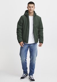 Grønn polstret jakke med hette, glidelås, og elastiske mansjetter, kombinert med blå jeans og svarte joggesko med hvite snøringer.