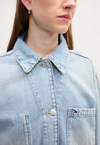 Veste en denim bleu clair avec un col classique, deux poches frontales, fermeture à boutons en métal argenté et détails de couture contrastants.