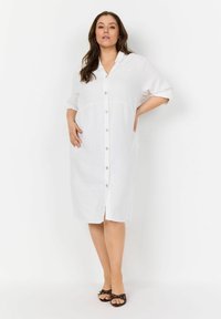 Weißes Leinenhemdkleid mit kurzen Ärmeln, kragen und einer Taillennaht. Kombiniert mit schwarzen Sandalen mit Riemchen.