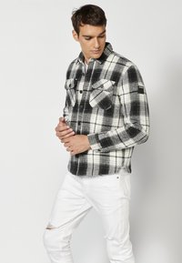 Zwart-wit checkered flanellen overhemd met twee borstzakken, knoopsluiting en lange mouwen; gedragen met verouderde witte jeans.
