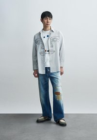 Světle šedá košile na knoflíky s kresbou obličeje, bílá tričko s barevnými puntíky, roztrhané modré džíny a boty s hnědými doplňky.