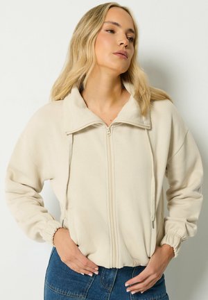 Lyshåret kvinde iført en beige fleecejakke med lynlås og blå jeans, stående med hænderne hvilende nær lommer mod en hvid baggrund.
