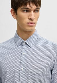 Camicia con bottone, caratterizzata da un motivo geometrico blu e bianco, colletto e maniche corte. Il tessuto appare liscio con un design a vestibilità regolare.