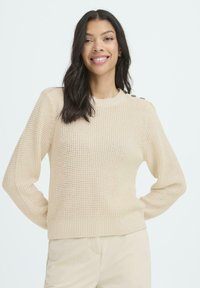 Mujer sonriente con cabello largo y oscuro, vistiendo un suéter de punto beige con botones en el hombro y pantalones beige a juego.