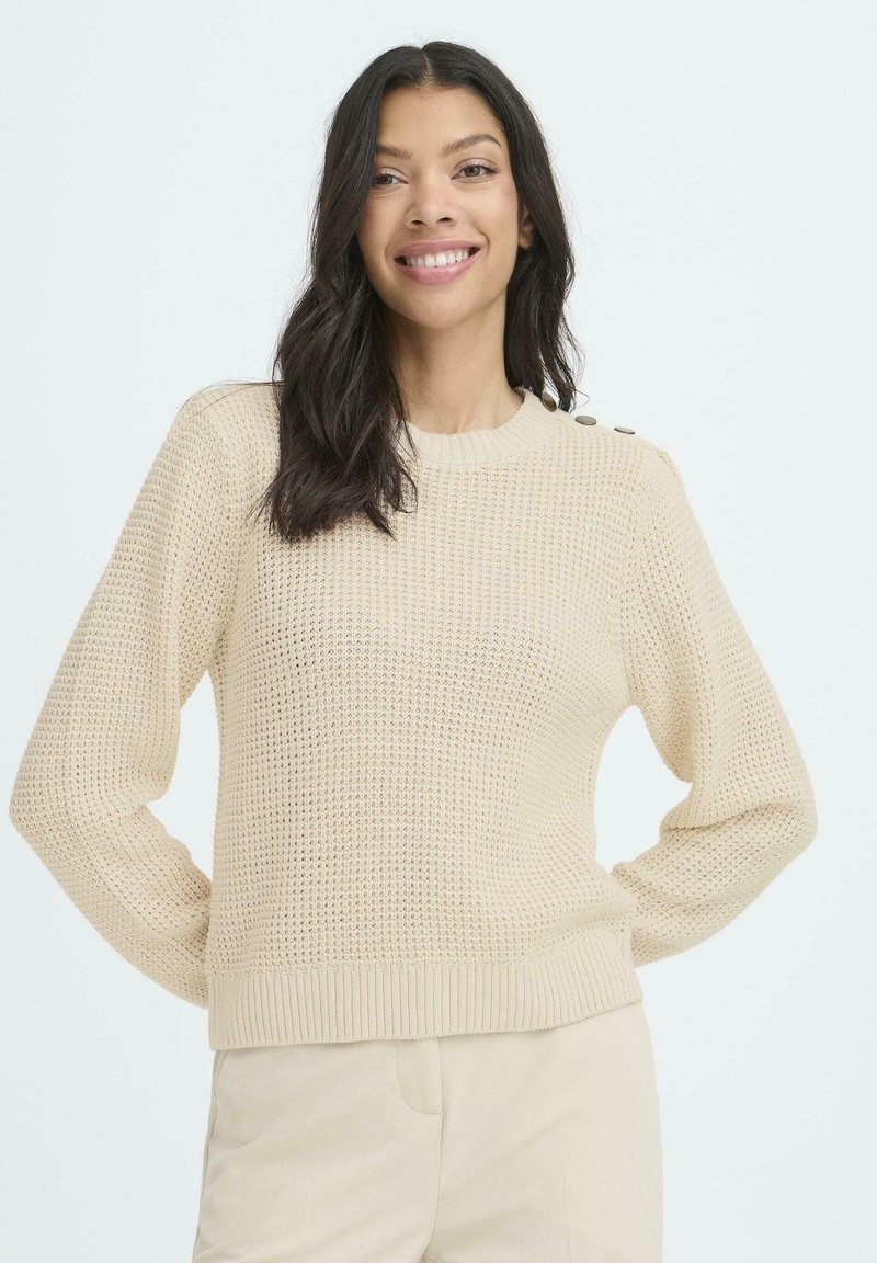 Mujer sonriente con cabello largo y oscuro, vistiendo un suéter de punto beige con botones en el hombro y pantalones beige a juego.