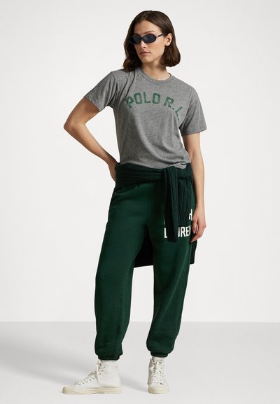 Pelēka kokvilnas t-krekls ar zaļu uzrakstu "POLO R.L.", kombinēts ar zaļiem sporta biksēm, uz kuriem ir baltais uzraksts "LAUREN". Modelis valkā saulesbrilles un sporta apavus.