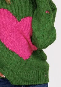 Groene gebreide trui met een grote roze hart op de voorkant, gedragen met blauwe jeans, hand in de taille gestoken.