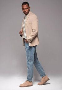 Blazer beige sobre una camisa a rayas, combinado con jeans de color azul claro y calzado beige. Textura suave, corte a medida y detalles mínimos.