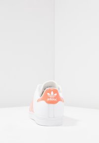 Vit Adidas-sneaker med korallfärgad hälflik och ränder, placerad med baksidan vänd mot oss på en vit yta mot en vit hörnvägg.