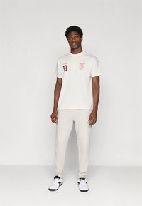 Muž stojící v bílém tričku s potiskem, světle šedých teplácích a bílých teniskách na jednobarevném světlém pozadí.