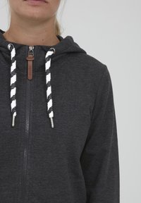 Mikina s kapucňou na zips v tmavosivej melírovanej látke. Obsahuje biele a čierne točené šnúrky, kovový zips a kožený akcent.