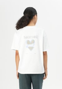 White cotton T-shirt with short sleeves, featuring "SAMSOE" text and a heart graphic in blue and beige on the back. Relaxed fit design.