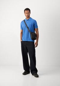 Blue polo shirt, black crossbody bag, dark loose-fitting pants, and black shoes. Smooth fabric, casual design, with a simple logo detail on the shirt.