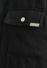 Veste en laine noire dotée d'une grande poche poitrine avec un fermoir à pression argenté et une étiquette de marque métallique, présentant une finition texturée et douce.
