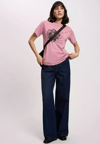 Różowa bawełniana koszulka z czarnym nadrukiem graficznym, zestawiona z ciemnoniebieskimi dżinsami o wysokim stanie i szerokich nogawkach. Niesie czarną torebkę na ramię.