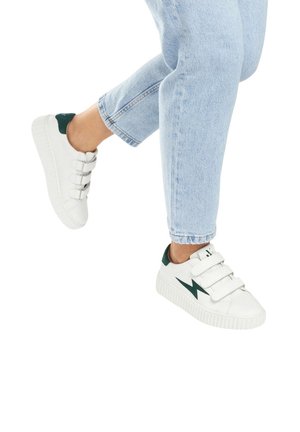 Des sneakers blanches avec des accents verts, trois bandes auto-agrippantes, une semelle texturée et un design d'éclair sur le côté. Portées avec un jean bleu clair.