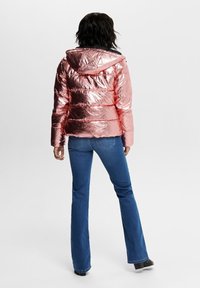 Rosa guld pufferjakke med en skinnende, tekstureret overflade og vandret syninger. Har en hætte og er parret med blå flared jeans.