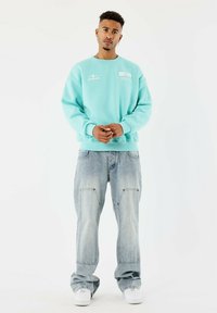 Un jeune homme portant un sweat à capuche turquoise, un jean bleu clair ample et des baskets blanches, se tenant avec les mains lâchement jointes.