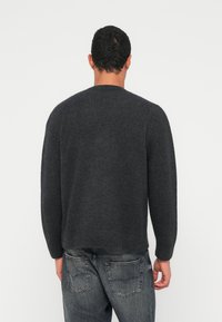 Άνδρας με την πλάτη, φοράει σκούρα γκρι πλεκτή μπλούζα και ξεθωριασμένο μαύρο τζιν μπροστά σε απλό λευκό φόντο.