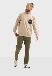 Sudadera de algodón beige con un bolsillo de parche negro, combinada con pantalones verde oliva y zapatillas blancas con acentos rojos y azules.